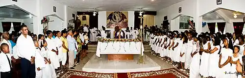 Afro-Dominicans attending catholic church service; with hands clasped in prayer and almost all dressed in white. During prayer in this picture they surround the alter and pray in unison.